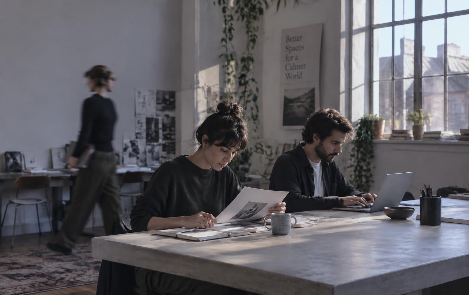 Two colleagues working quietly at a studio table, one reading a printed sheet while the other types